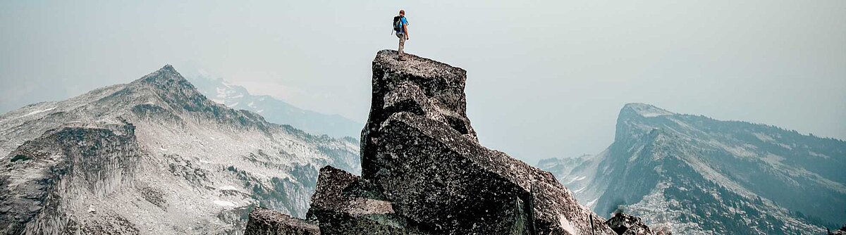 Eine Person steht auf einem Berggipfel und und blickt in die Ferne. Im Hintergrund sind weitere schneebedeckte Berge zu sehen.