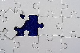 Bild zeigt eine Fläche mit weißen Puzzleteilen. Auf dieser Flächs fehlt ein Teil, welches bereits bereit liegt - d.h. das Puzzle ist fast fertig.